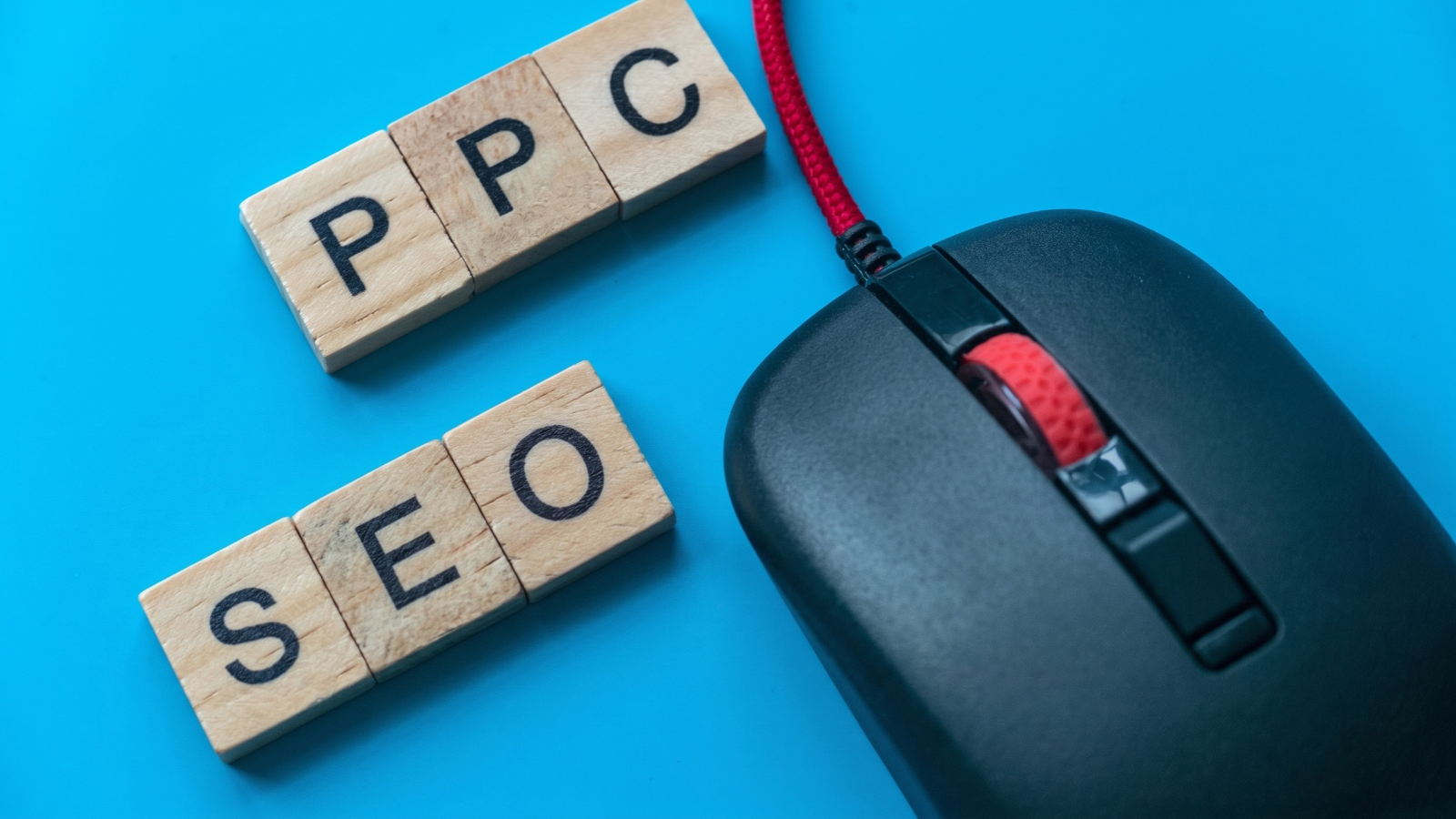 Wooden tiles spelling "PPC" and "SEO" next to a black and red computer mouse on a blue surface.