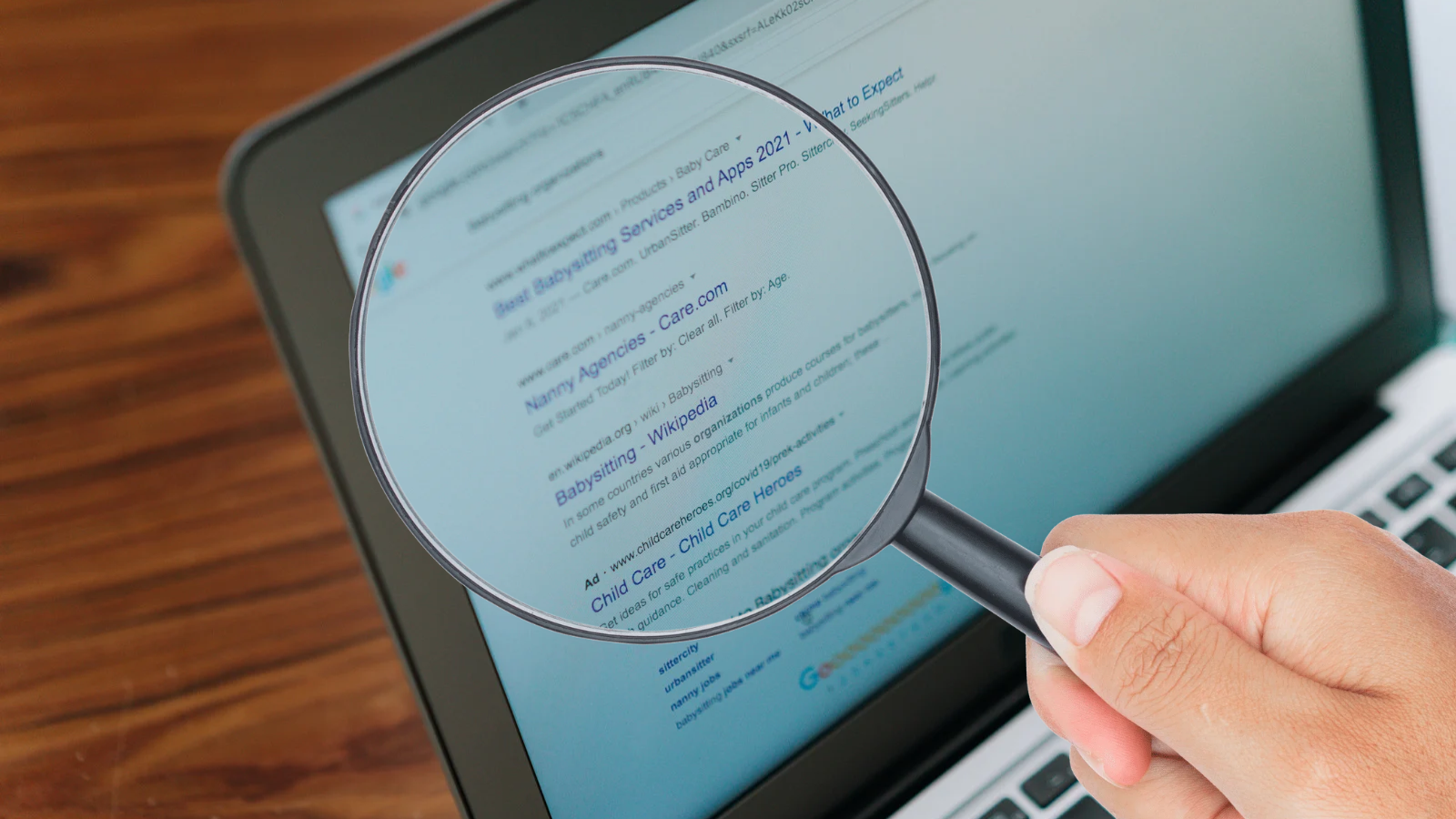 Hand holding a magnifying glass over a laptop screen displaying search engine results.